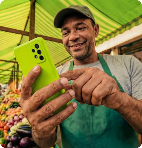 Feirante sorrindo com o celular na mão, na banca de frutas